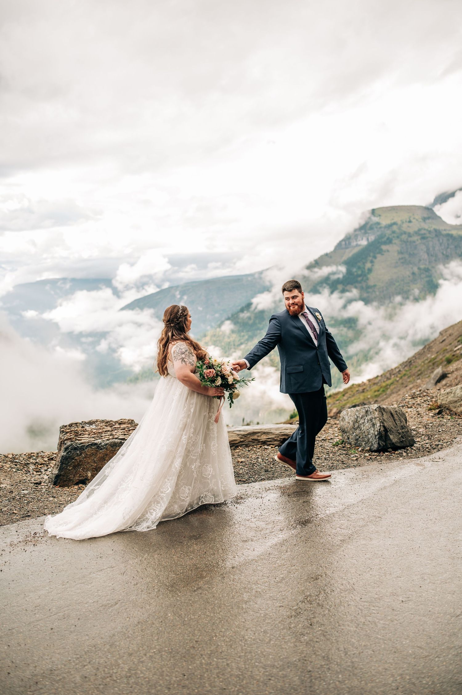 A couple in wedding attire playfully poses on a misty mountain road with dramatic clouds and peaks in the background.
