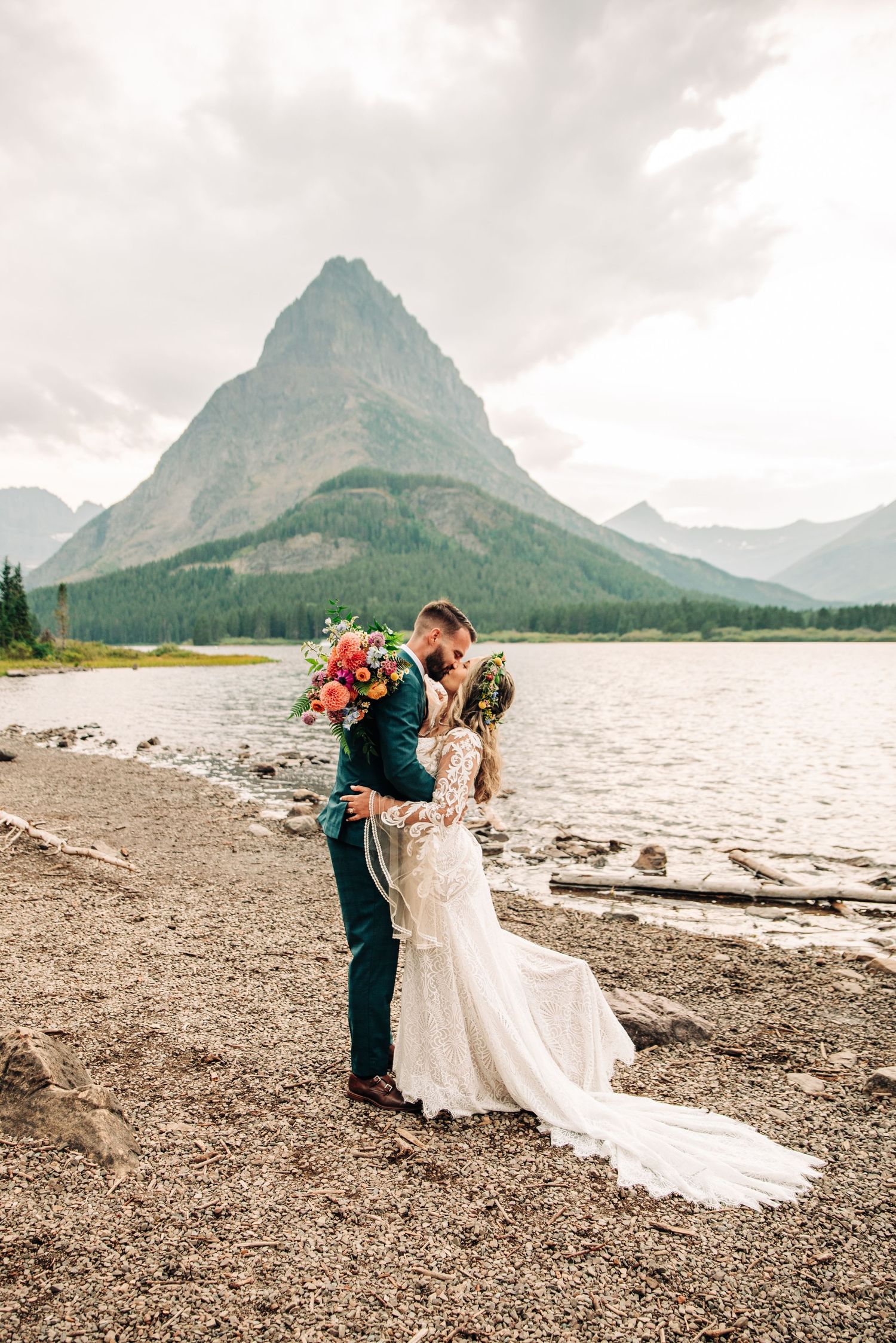 A couple embraces by lake shore with colorful bouquet and majestic mountain peak rising in background at Glacier National Park.