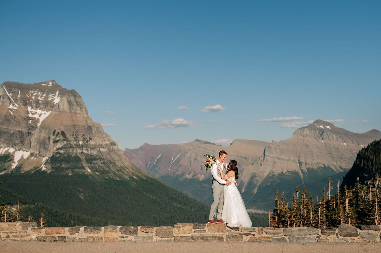 A couple embraces on a scenic mountain overlook at Glacier National Park with snow-capped peaks in the background.