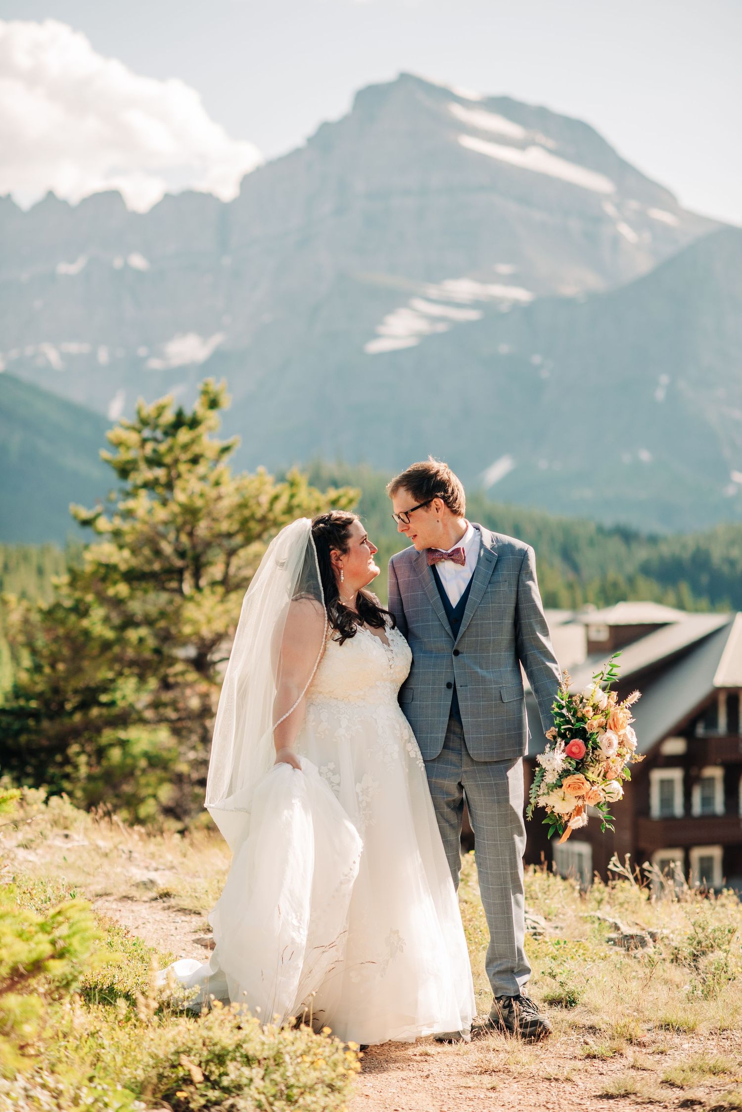 Wedding couple poses against majestic mountain backdrop with rustic lodge in background.