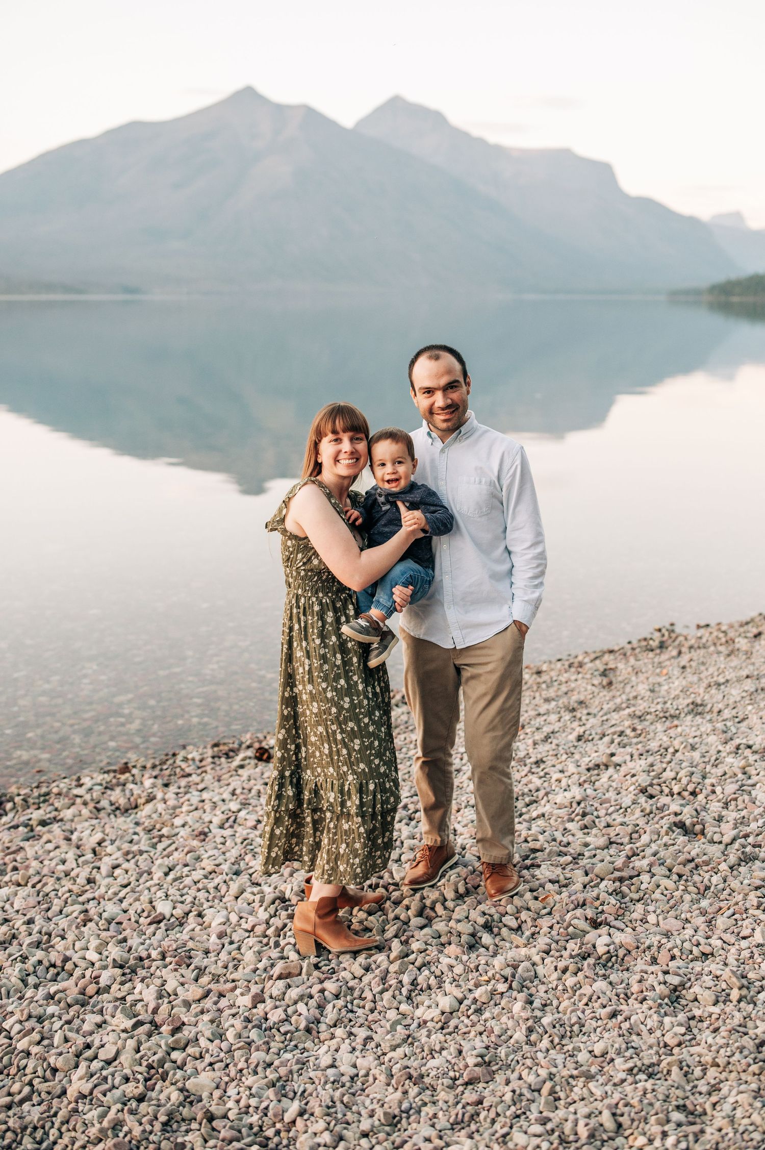 Family stands on rocky lakeshore with mountain peaks and reflective waters providing scenic backdrop in nature.