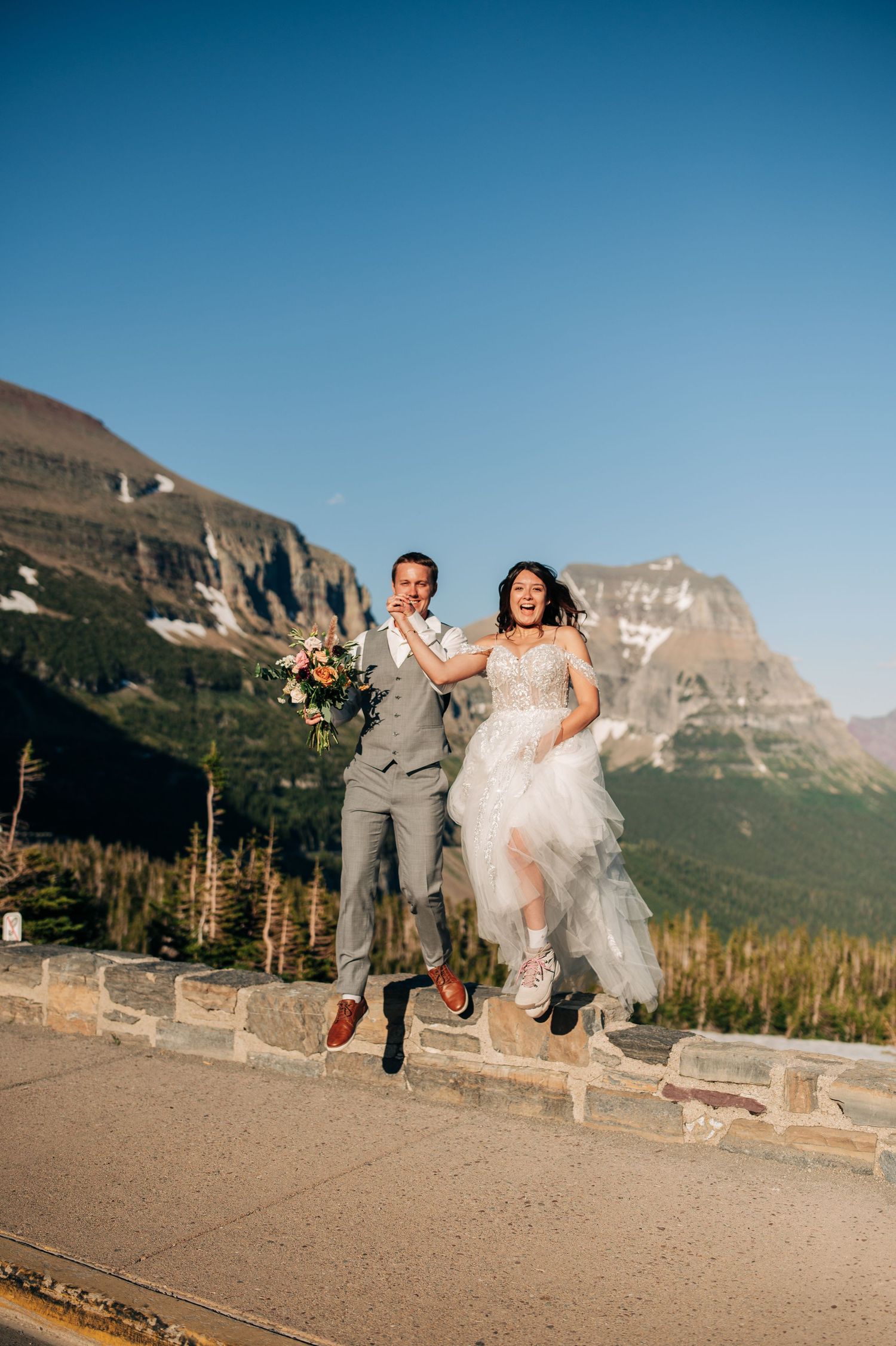 Two people in wedding attire joyfully jumping in the air with majestic mountains and blue sky at Glacier National Park.