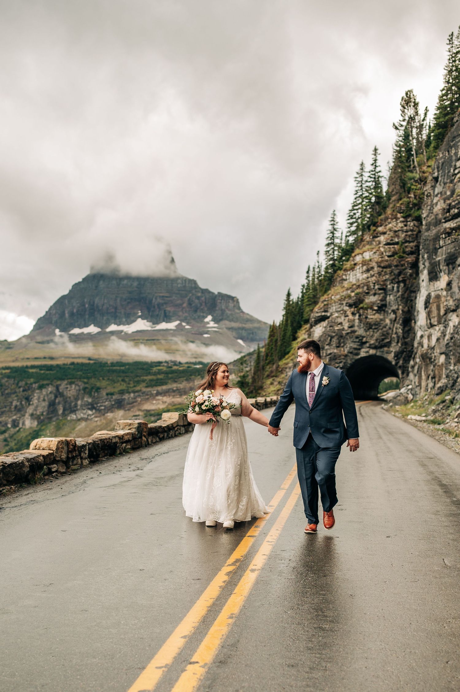 Couple walking along scenic mountain road at Going-to-the-Sun Road in Glacier National Park on cloudy day.