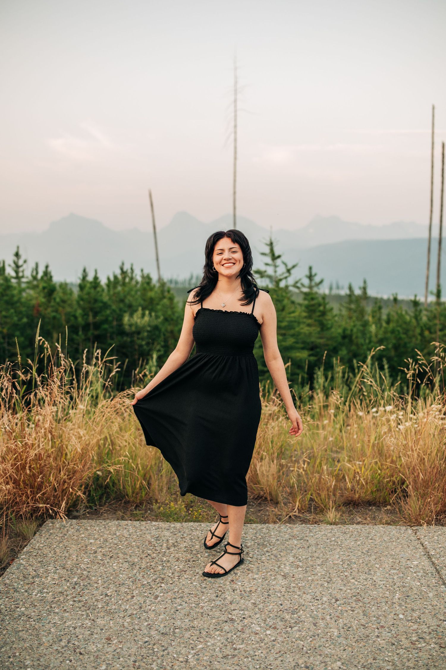 Person in a black summer dress poses on a path with mountains and evergreen forest in the misty background.
