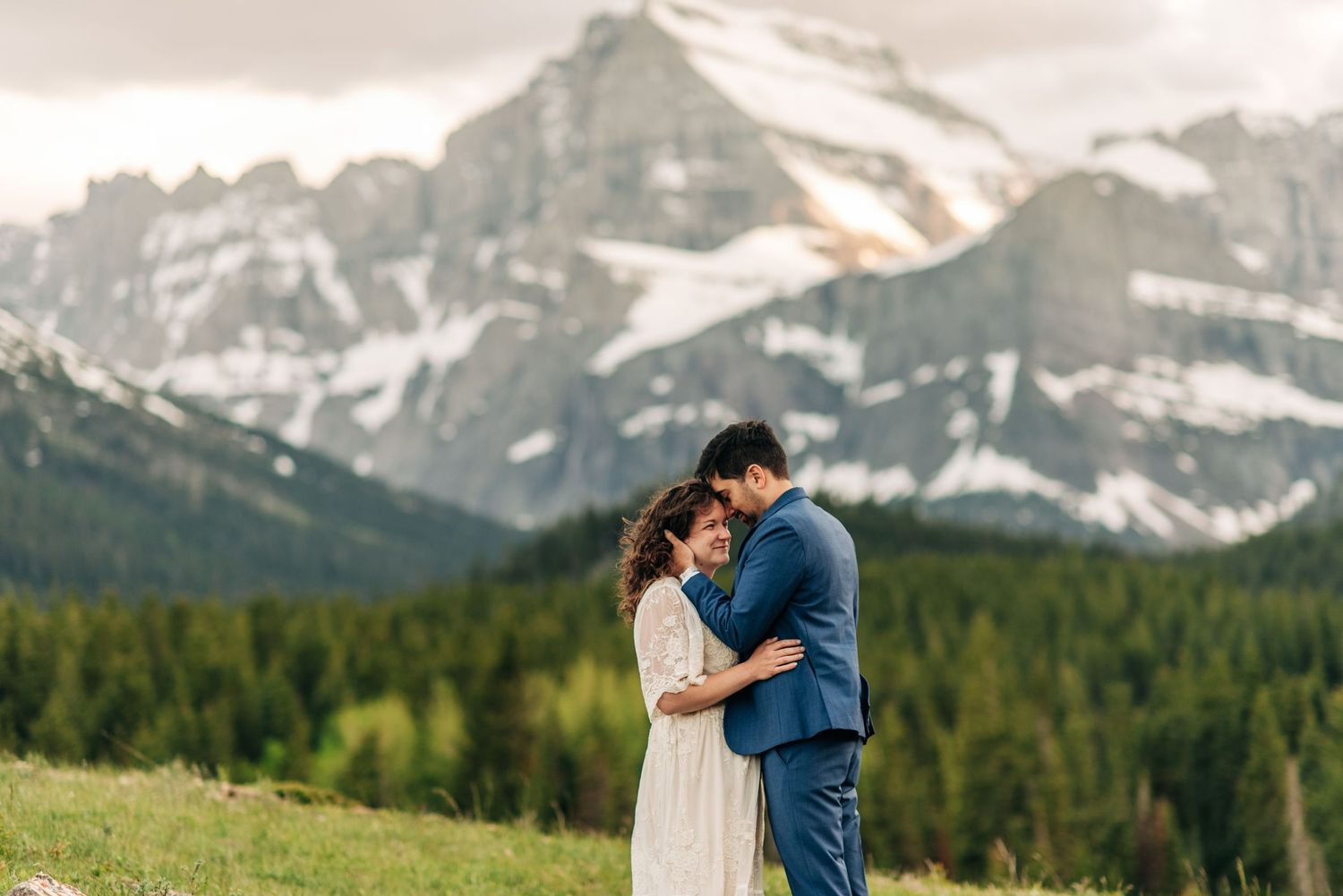 A couple shares an intimate moment while standing together in front of snow-capped mountain peaks and evergreen forest.