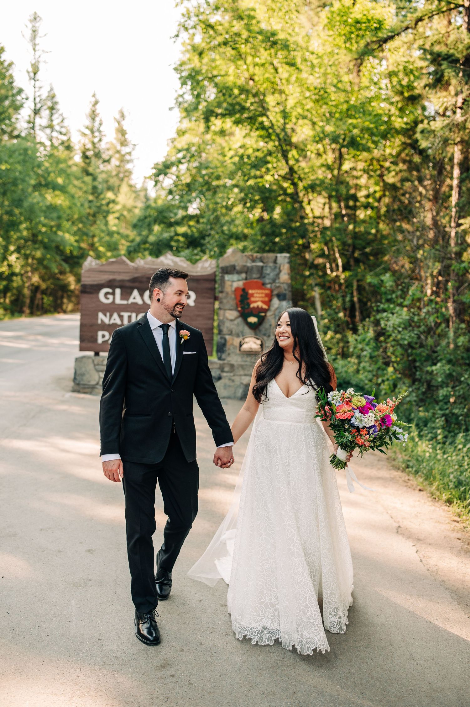 couple holding hands walking towards the camera in front of the glacier national park entrance sign