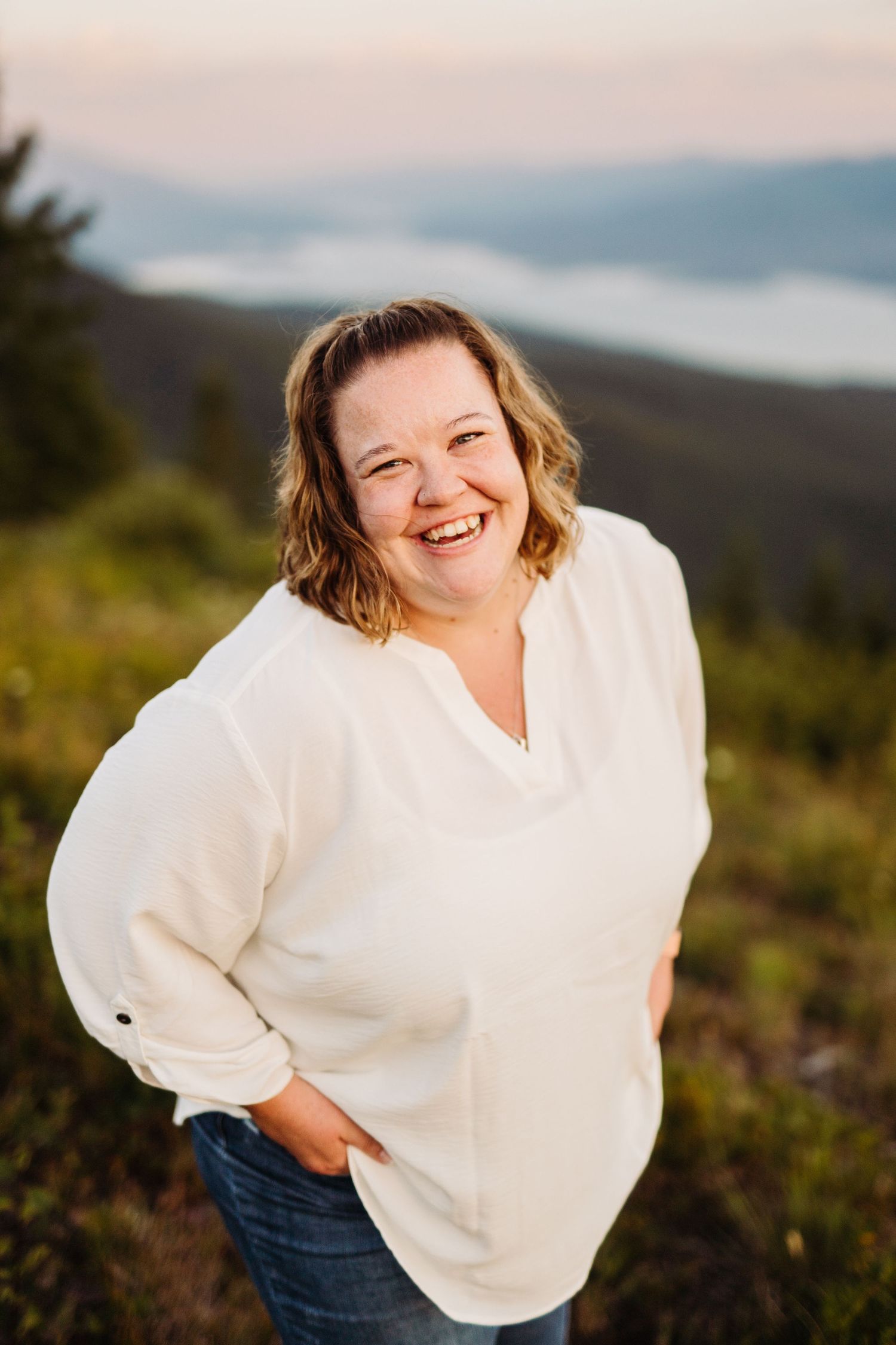 A person in a white sweater smiles warmly while standing on a hillside with mountains in the background at sunset.