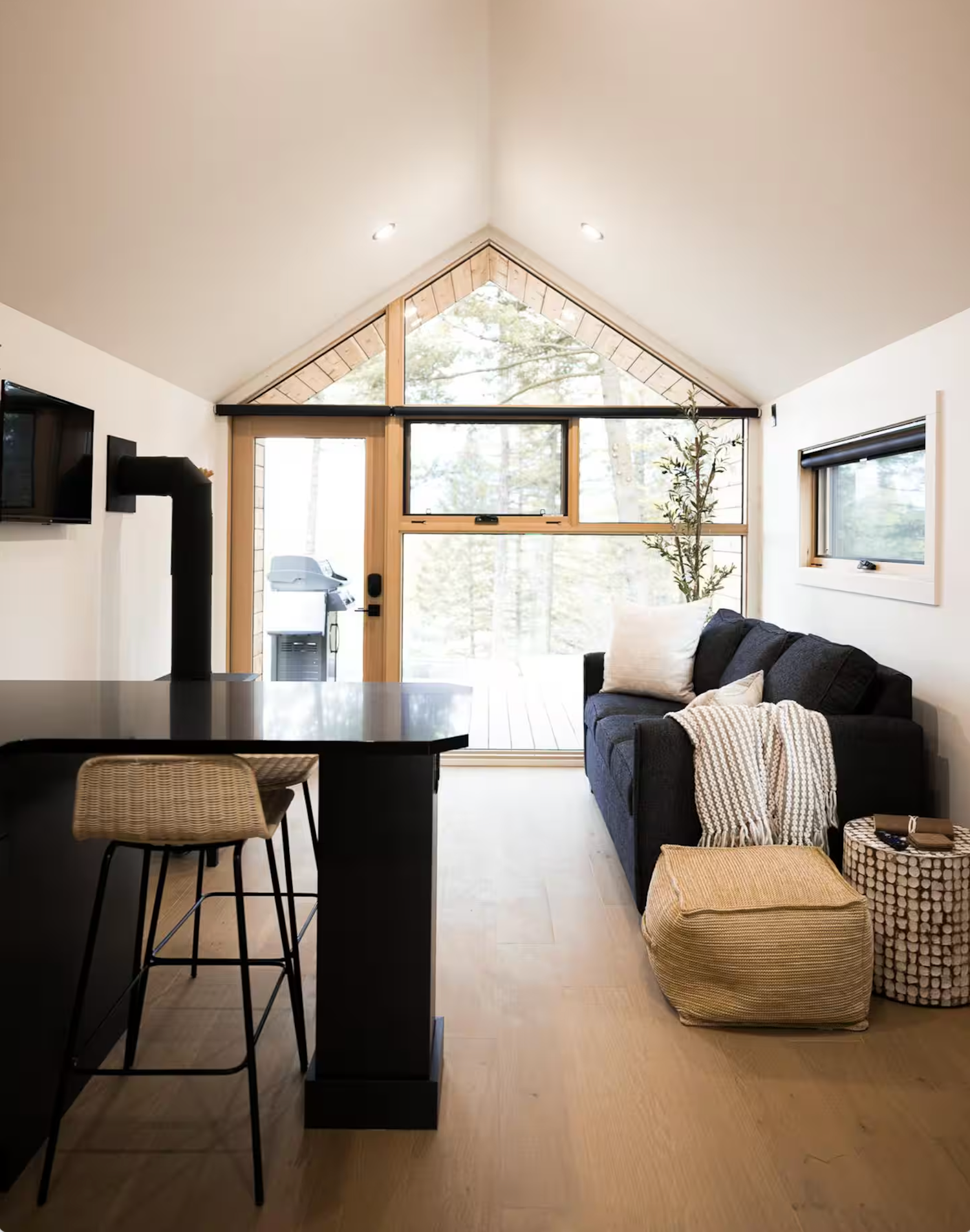 Modern tiny house interior with vaulted ceiling, black sofa, kitchen island and large triangular window overlooking nature.