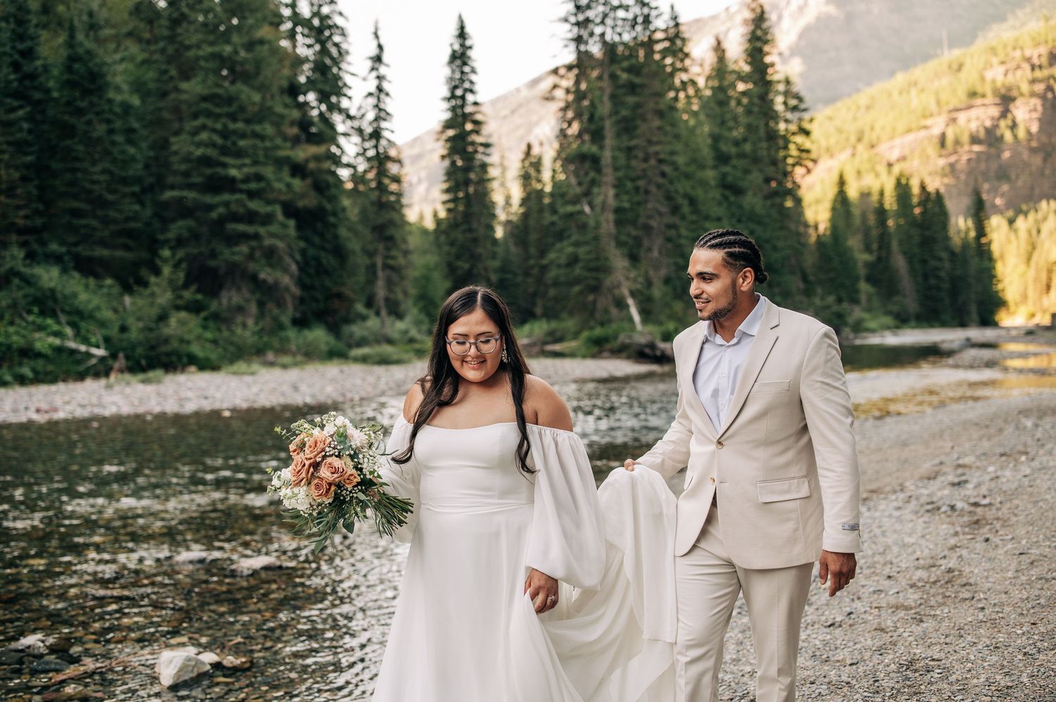 Two people in white outfits walk along a mountain trail with evergreen trees and golden sunlight in the background.