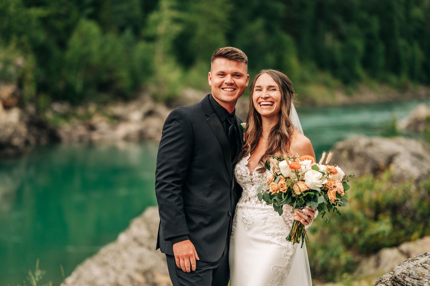 A couple poses together near a serene turquoise lake with a scenic mountain backdrop during wedding photos.