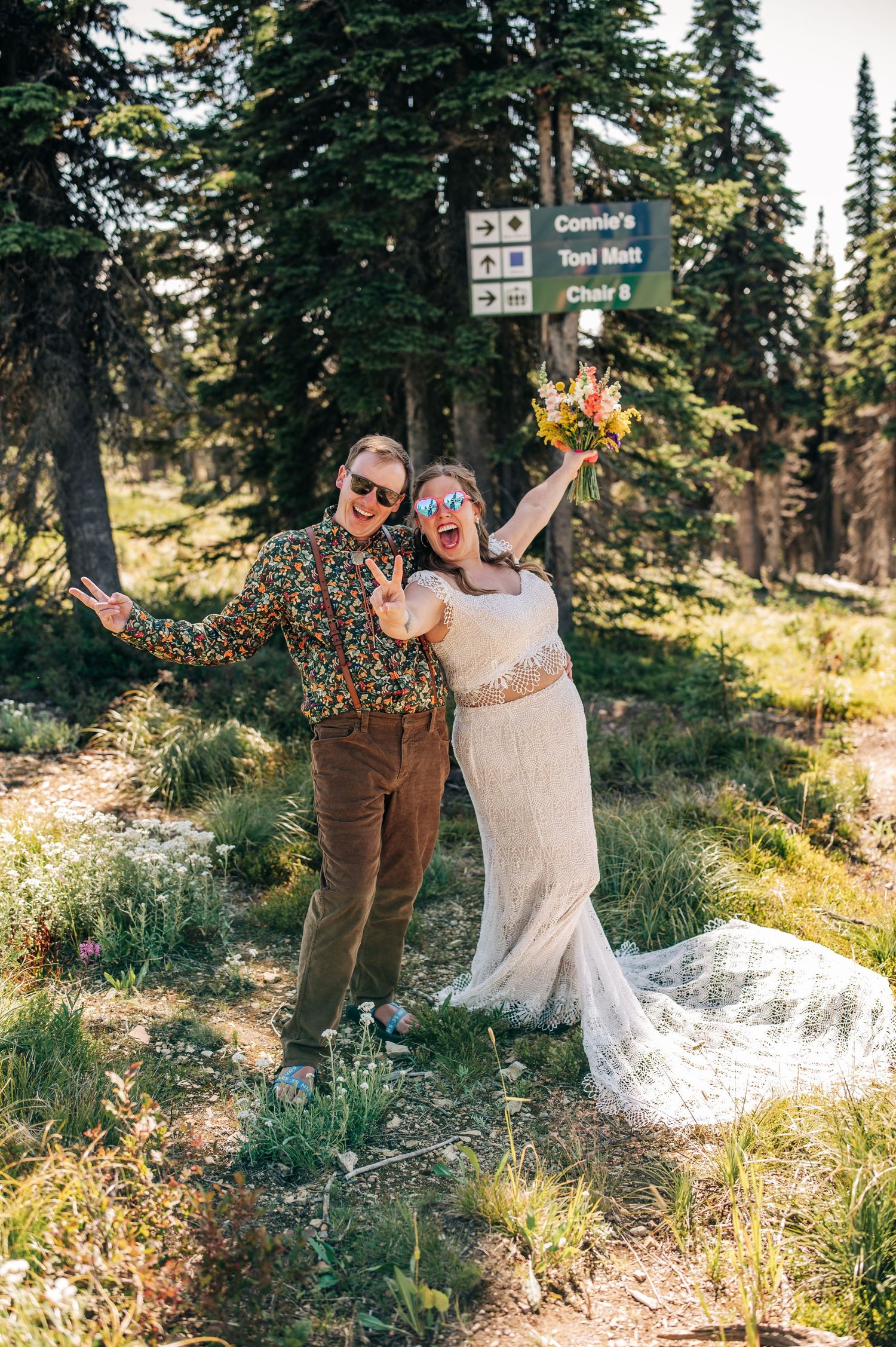 Joyful couple in wedding attire posing at mountain trail sign surrounded by pine trees on their elopement day.