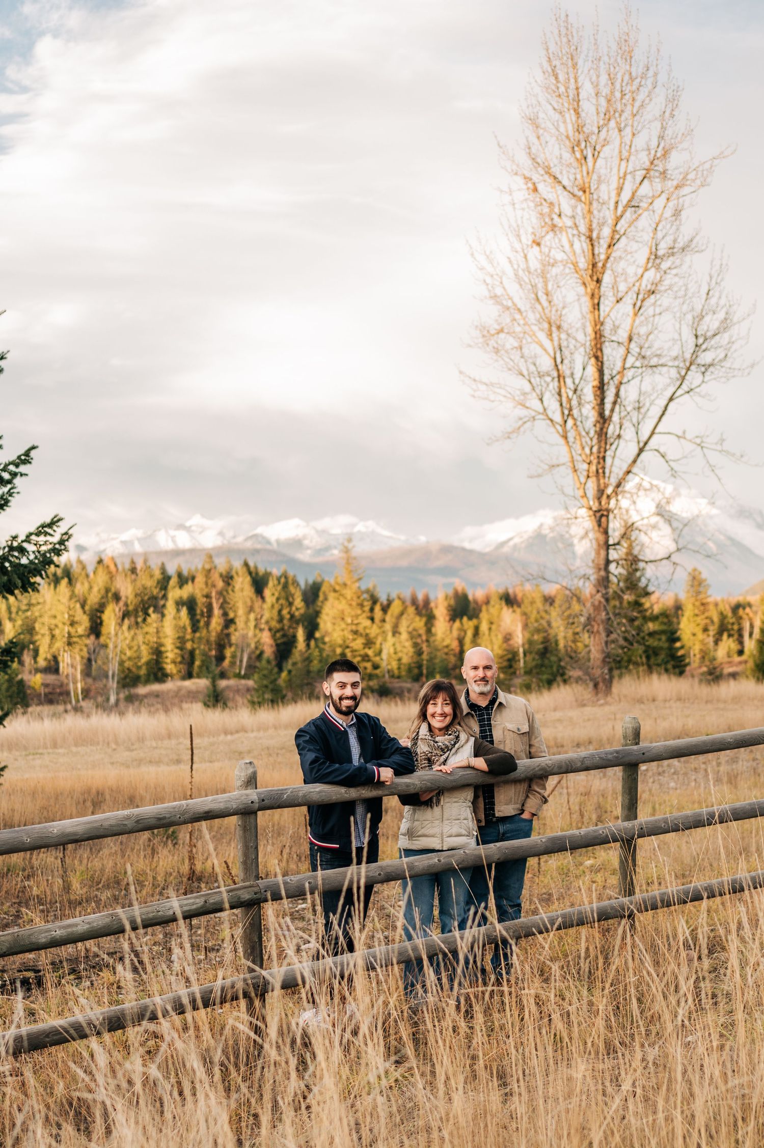 Three people stand at a wooden fence with snow-capped mountains and autumn trees in the background on a cloudy day.