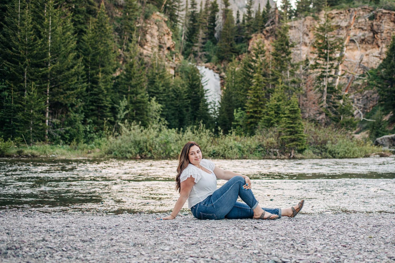 Someone relaxes on a rocky riverbank surrounded by evergreen forest and rocky cliffs in a scenic mountain landscape.
