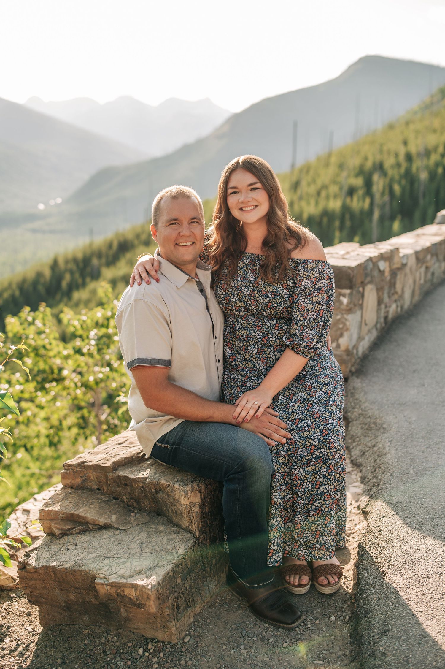 Two people sit together on a stone wall overlooking scenic mountain views on a sunny summer day.