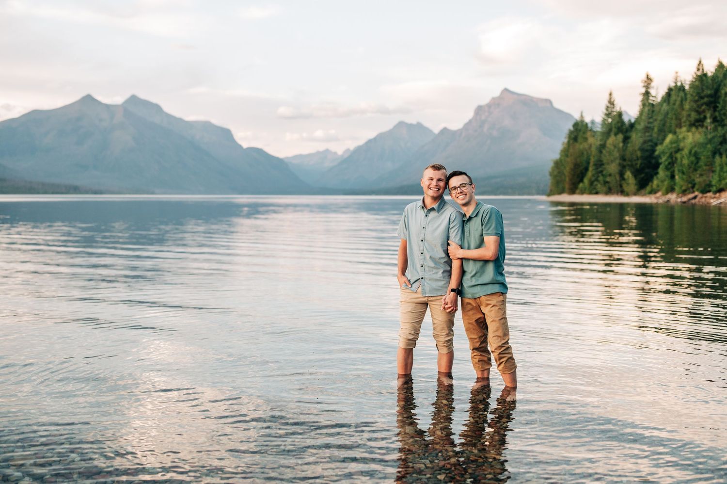 Two people standing in shallow water at sunset with mountains and forest reflecting in a serene lake.