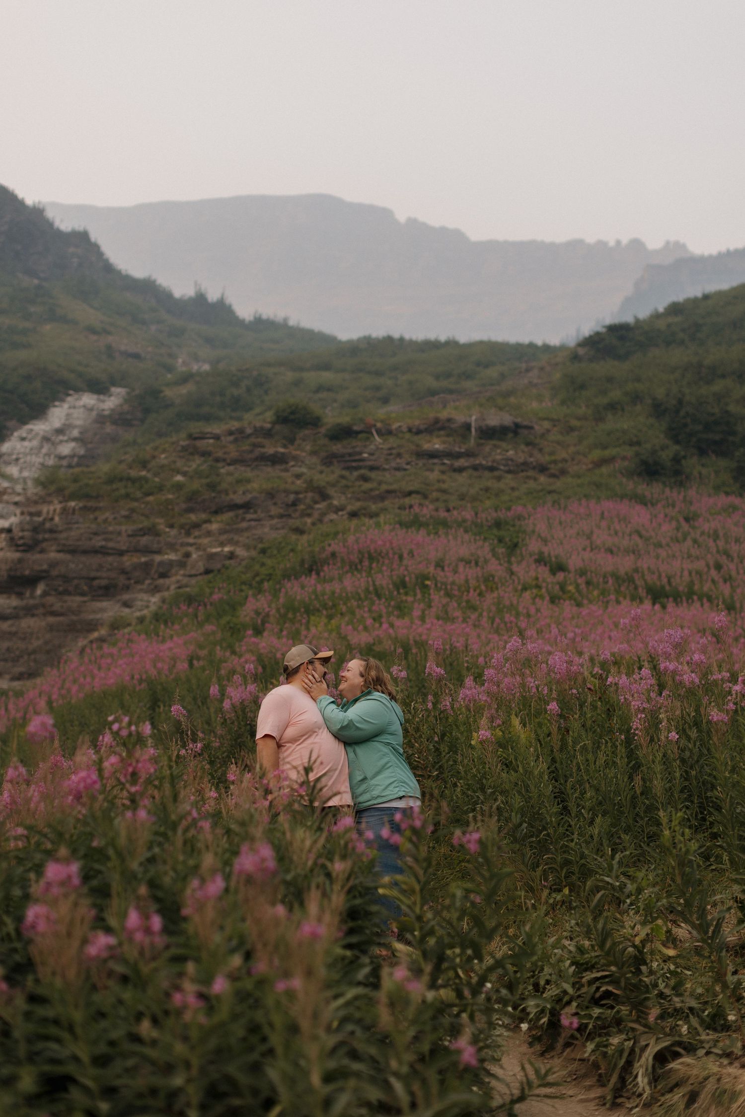 Couple embracing romantically in a field of pink wildflowers with misty mountain landscape in the background.