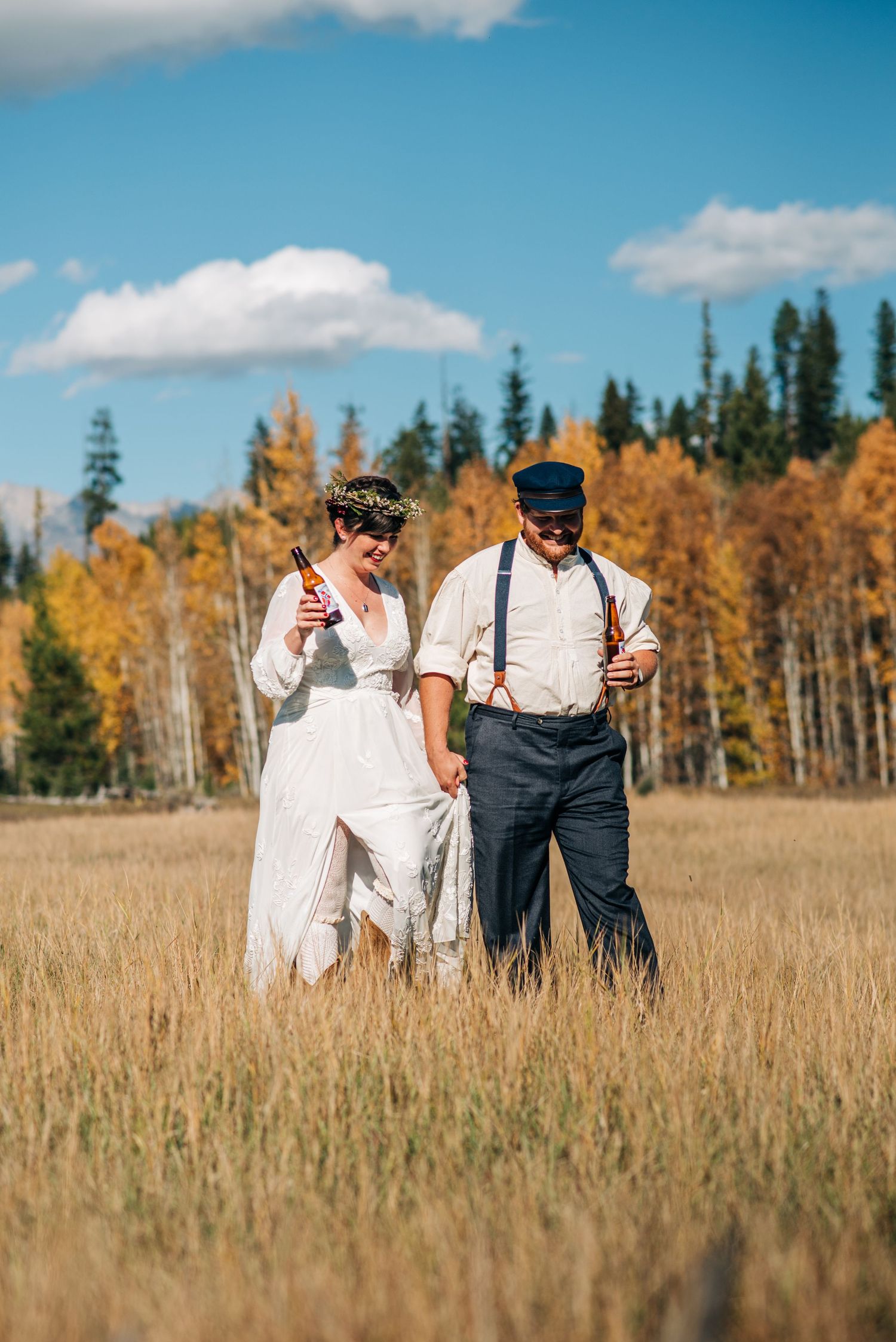 Two people in vintage-inspired attire walk through a golden autumn field with evergreen trees and blue sky in background.