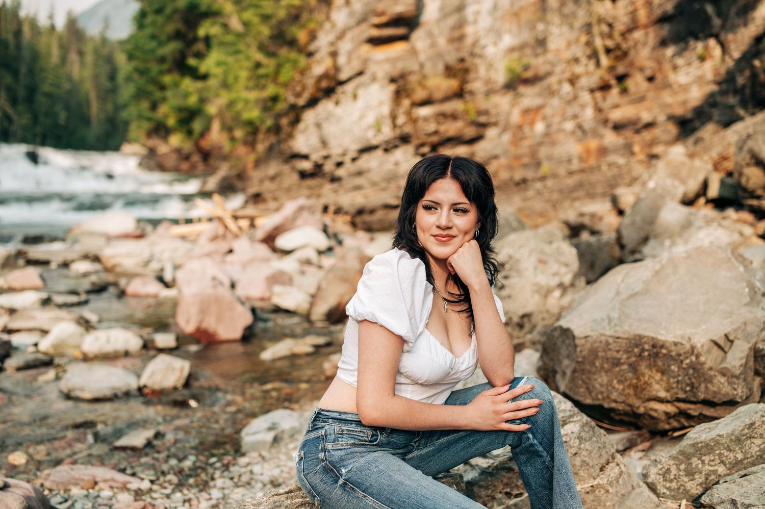 Someone sits casually on rocky riverbank with rushing water and cliffs in scenic mountain wilderness landscape.