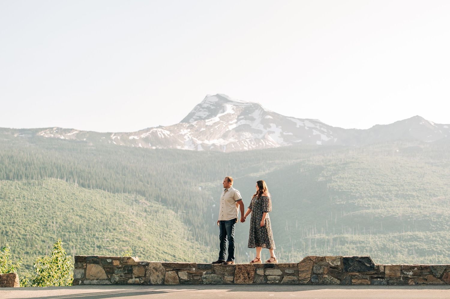 Two people walk along a stone wall overlooking scenic mountain views in a nature setting with a green valley below.