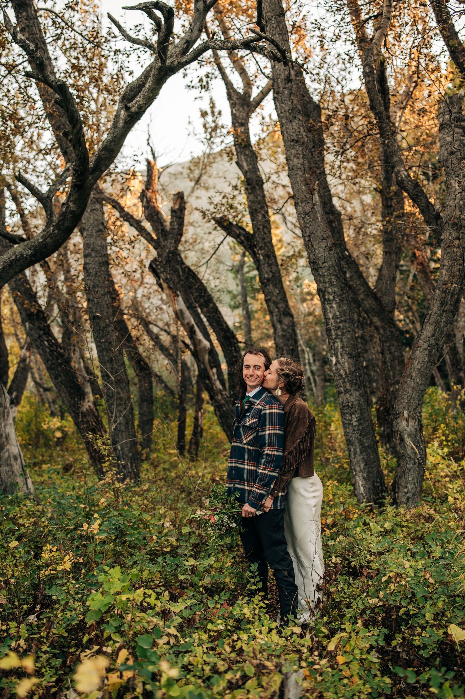 A couple embraces among autumn trees in a wooded setting with golden leaves and natural light filtering through branches.