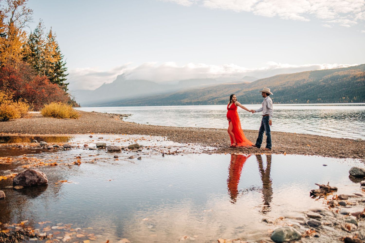 A couple stands on a rocky lakeshore at sunrise with mountains and autumn trees in the background.
