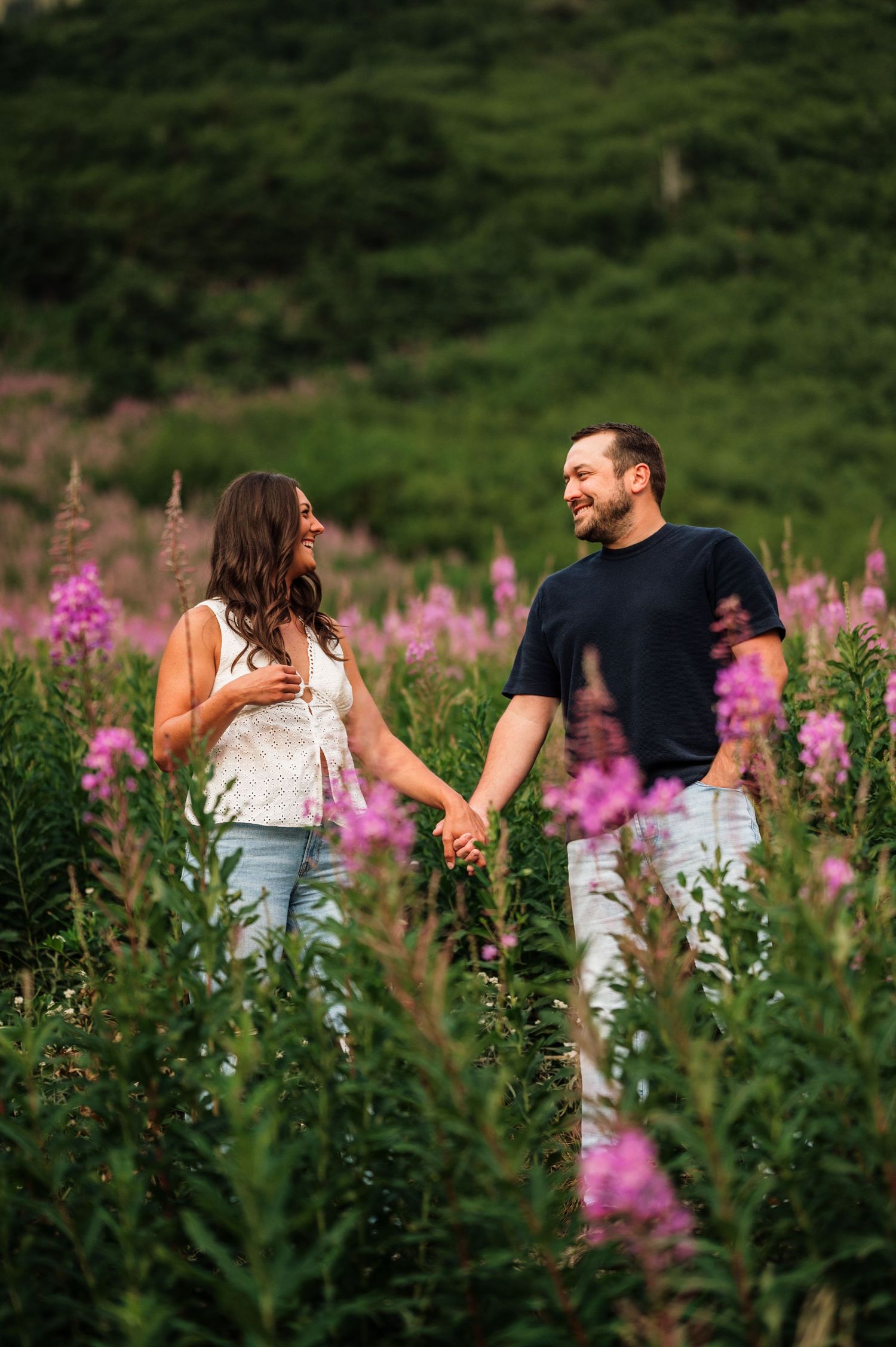 Couple holding hands and smiling in a field of pink wildflowers with lush green hills in the background.