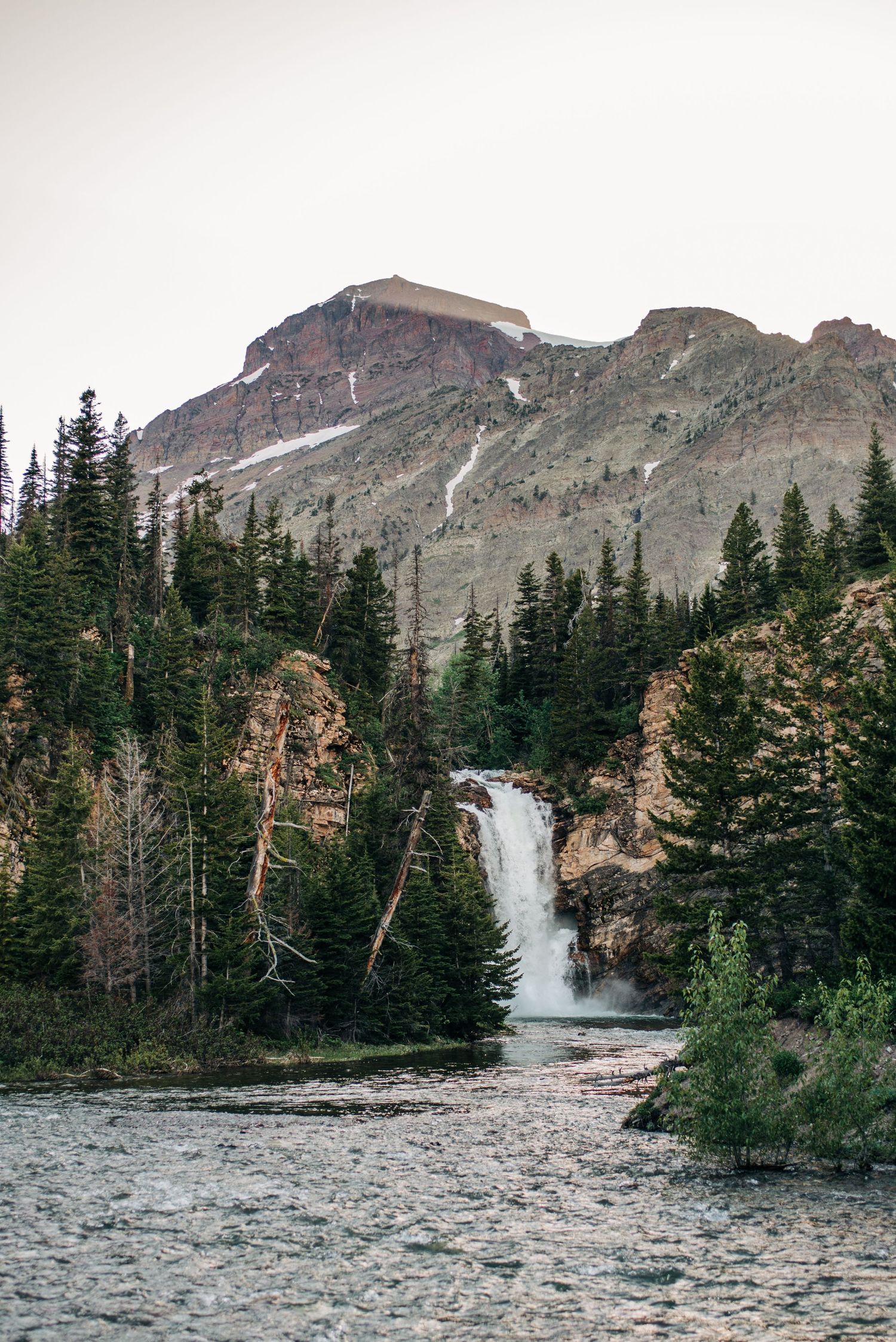 A scenic mountain waterfall cascades through evergreen forest surrounded by rocky terrain and patches of snow.