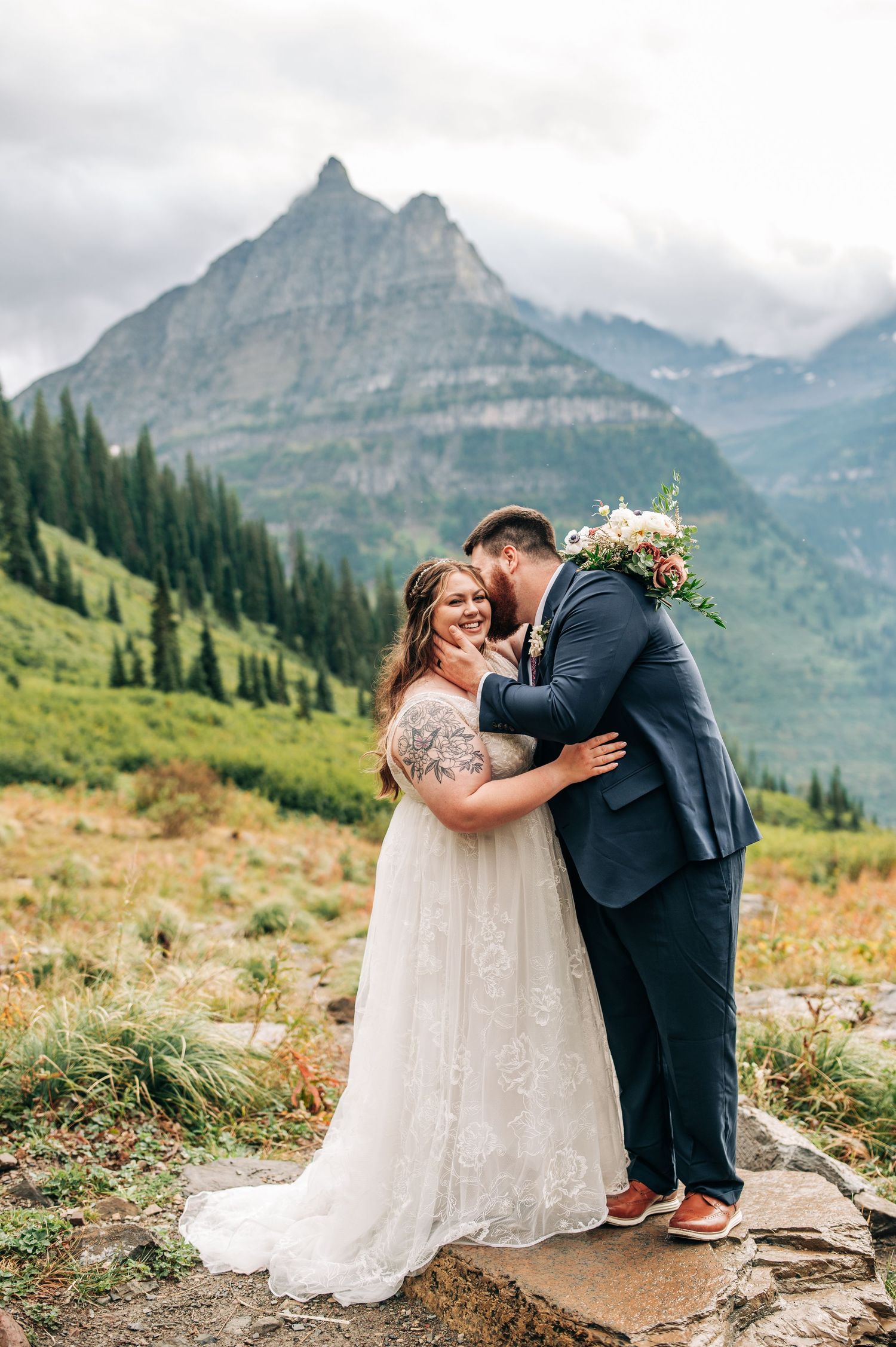 A couple shares an intimate moment while standing on a rocky outcrop with majestic mountain peaks and pine forests in the background.