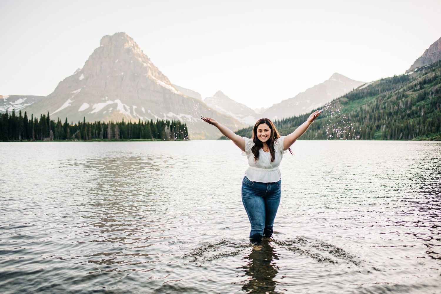 Person standing in lake water with arms raised joyfully against mountain backdrop at Two Medicine Lake in Glacier National Park.