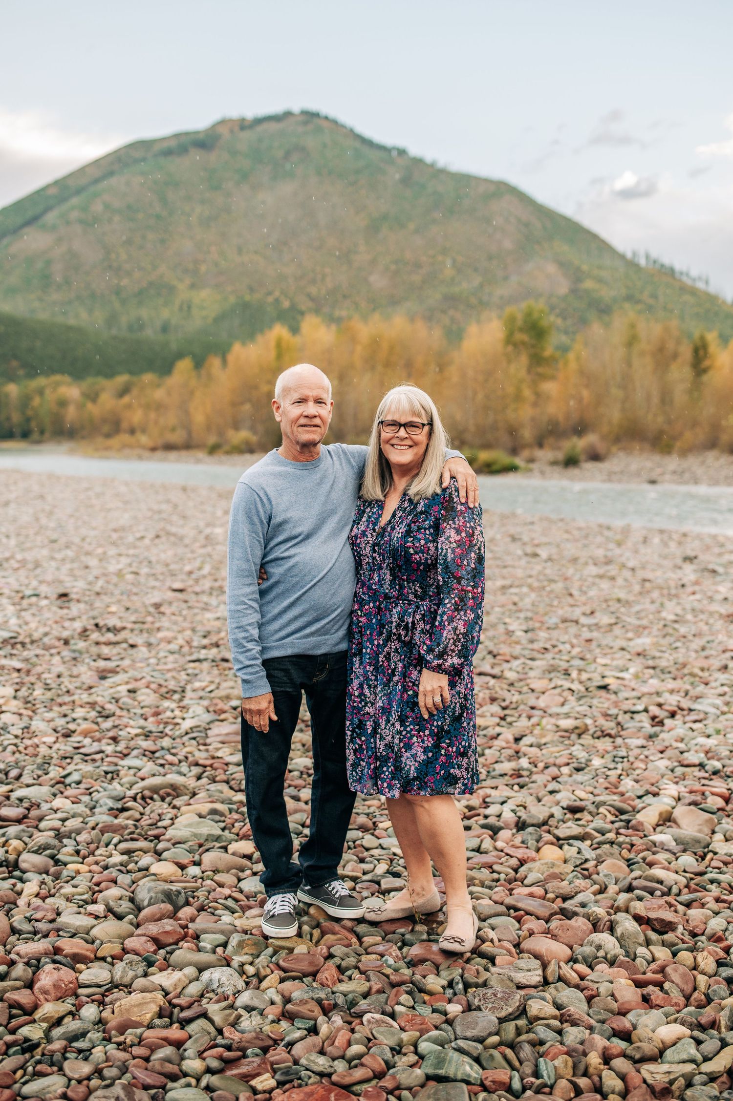 A couple stands together on a rocky riverbank with autumn trees and a mountain in the background.