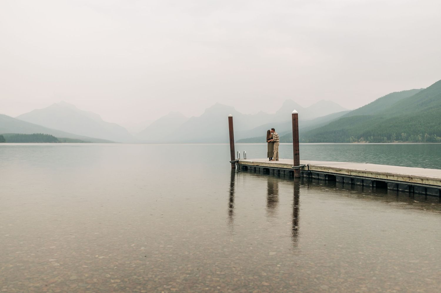 A solitary figure stands on a misty wooden pier extending into a serene mountain lake on an overcast day.
