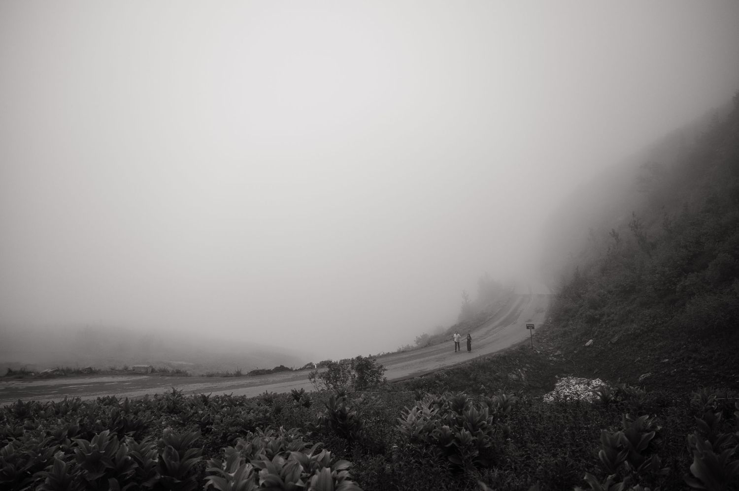 Black and white photo of a foggy mountain road with two distant figures walking along the misty curved path.