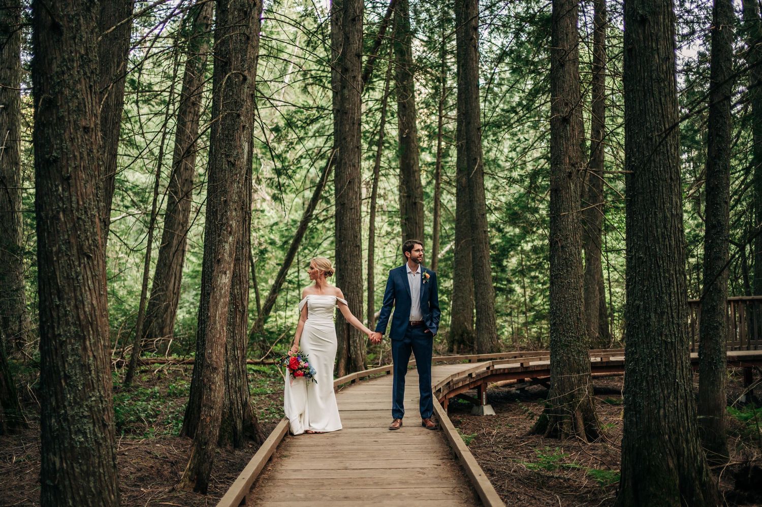 A wedding couple walks hand in hand down a serene forest path lined with tall evergreen trees in a natural setting.