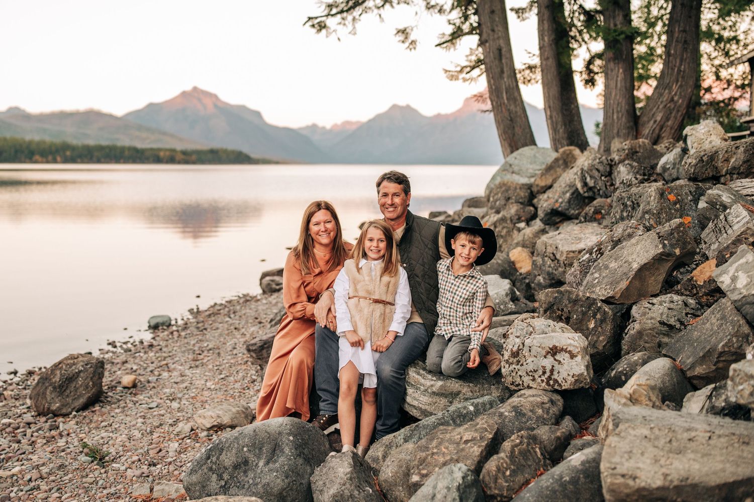 A group sits together on rocky shoreline at sunset with mountain lake and trees in background.