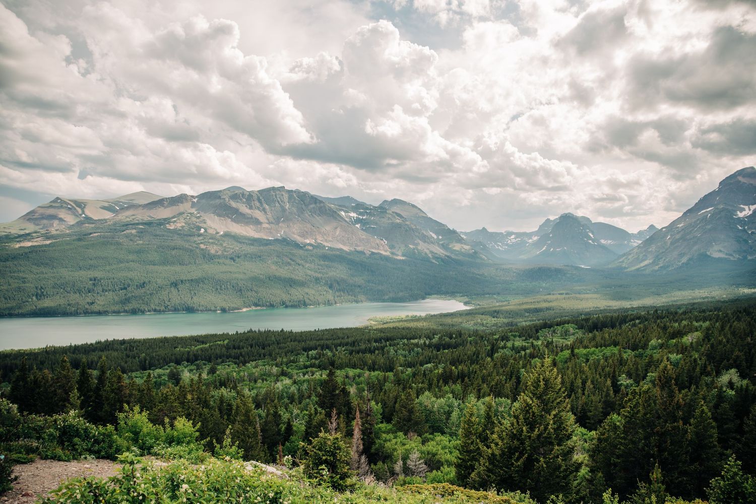 A dramatic mountain landscape with a lake nestled between peaks and lush green forest in the foreground under cloudy skies.