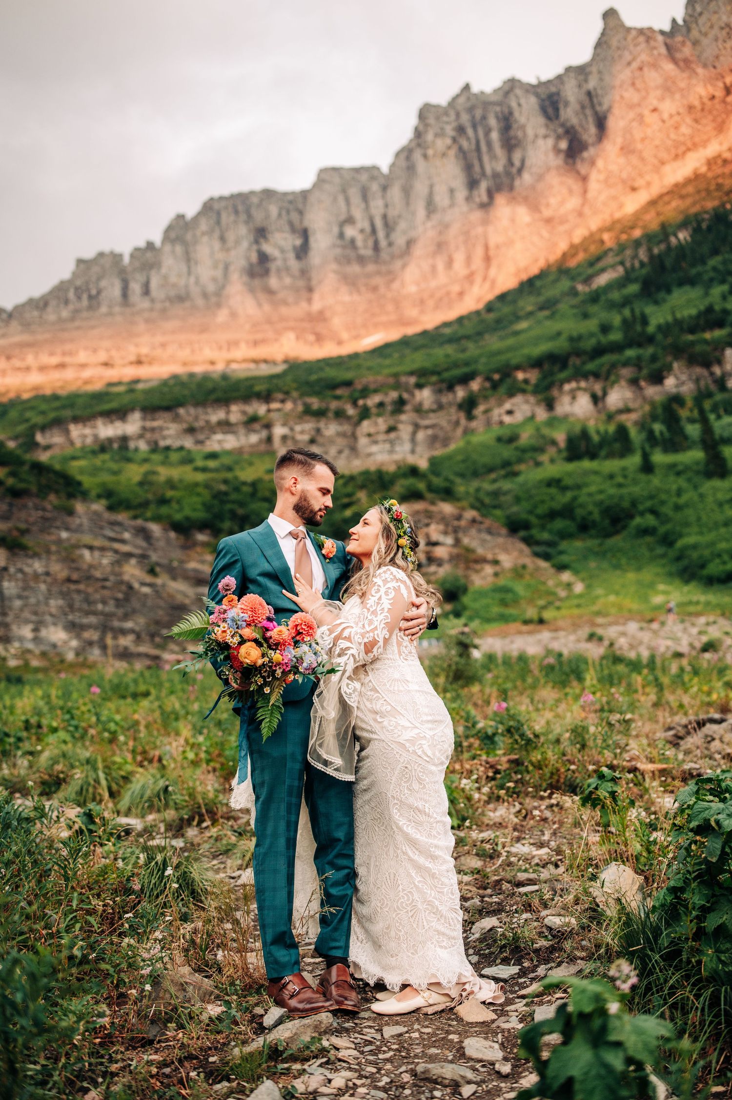 Two people embrace in a romantic moment against a dramatic mountain landscape during sunset.