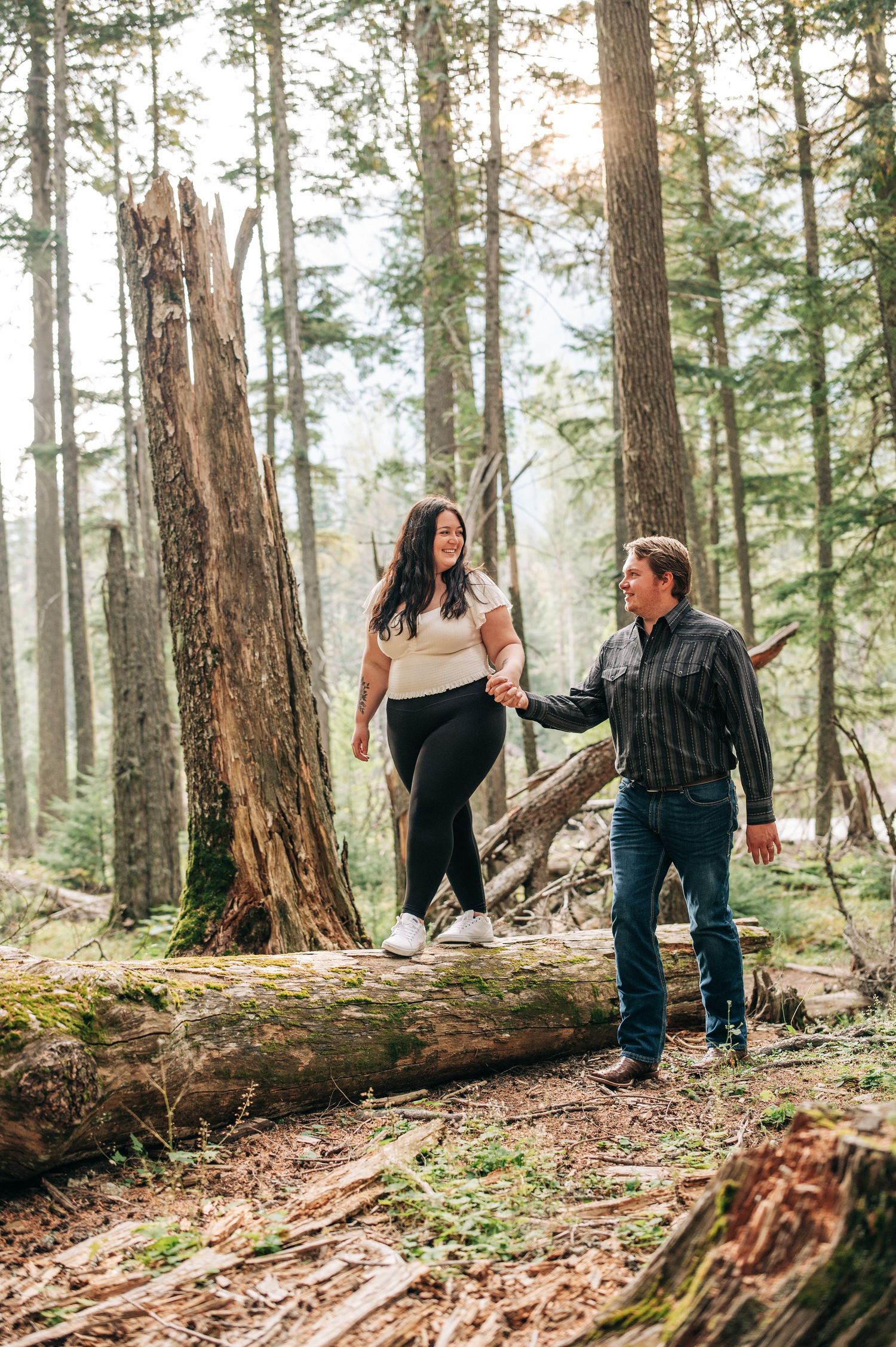 Two people holding hands while balancing on a fallen log in a misty evergreen forest.