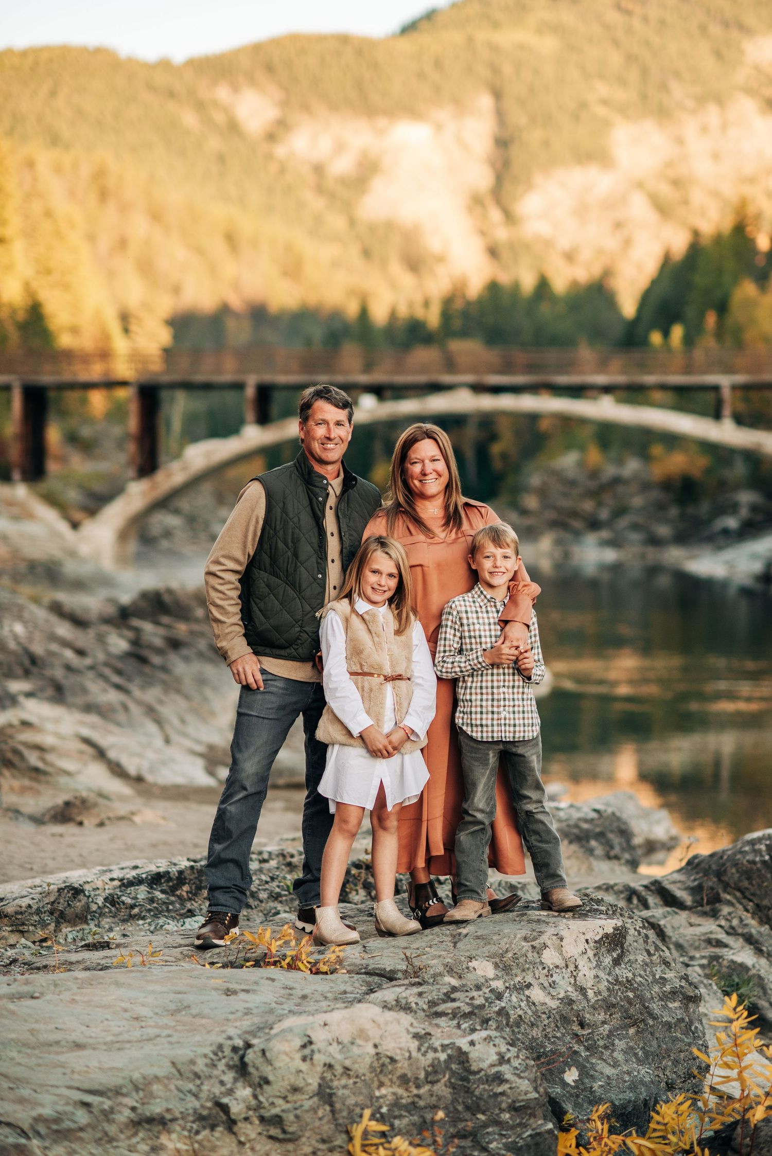 Family stands together on rocky riverbank with scenic arched bridge and golden hills in background during sunset.