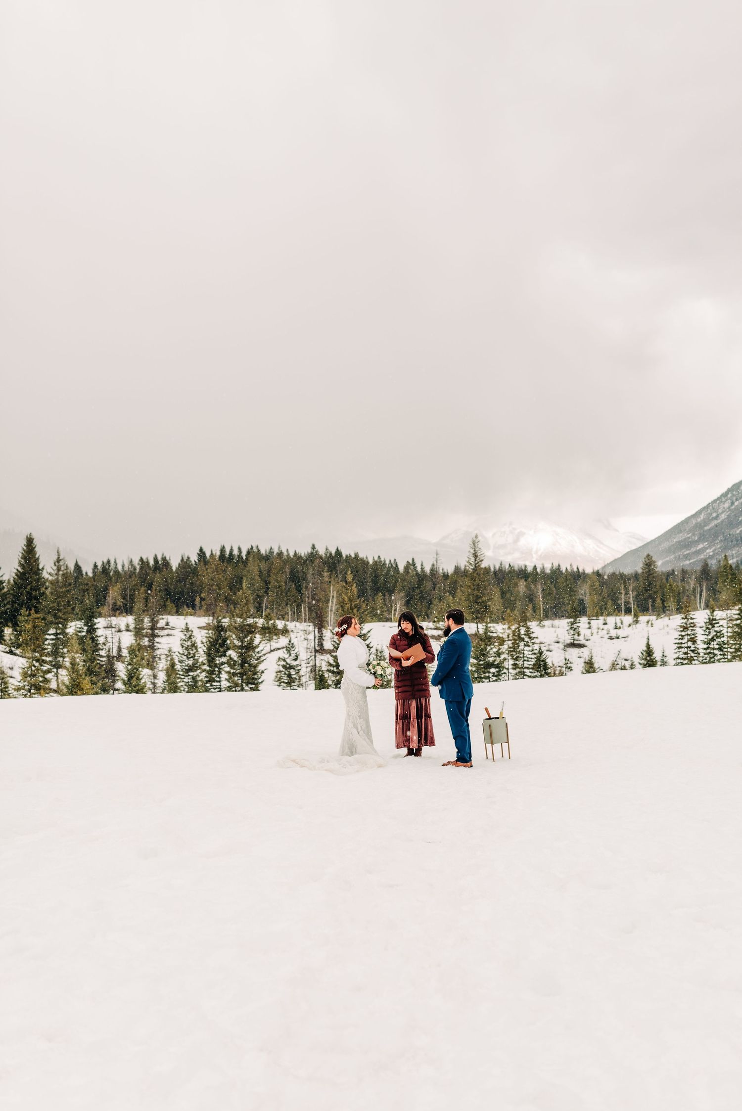 Three people standing in a snowy mountain valley surrounded by evergreen forest.