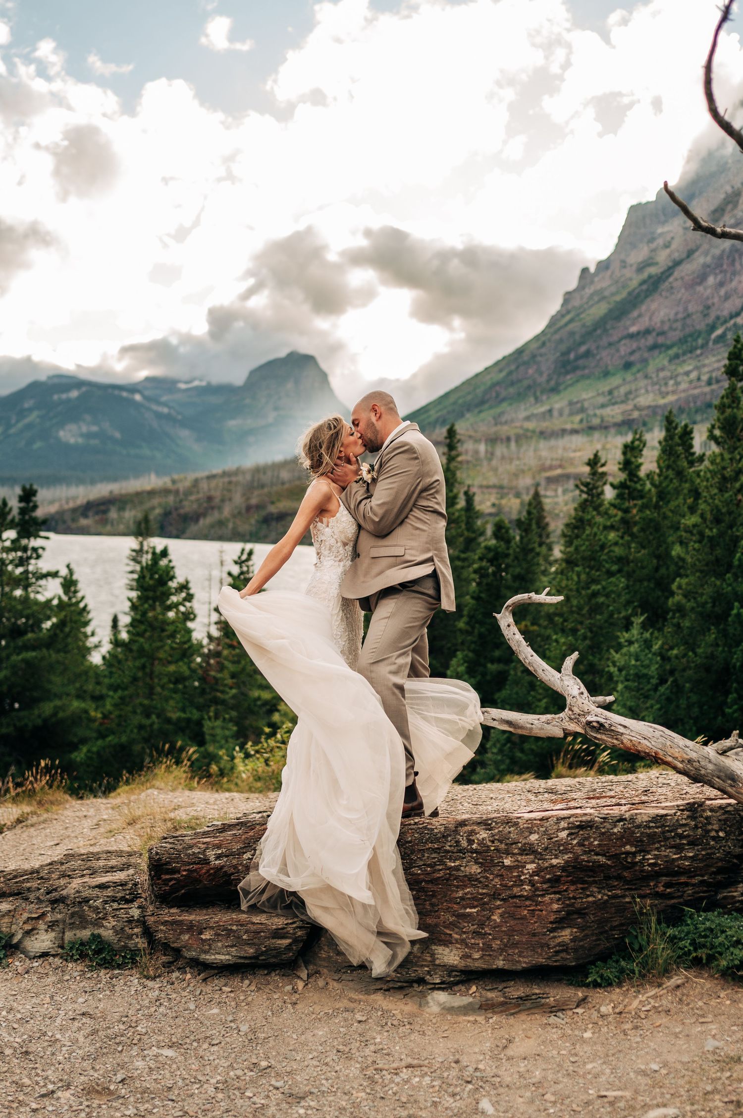 A couple in wedding attire poses playfully on a fallen log with majestic mountain peaks and cloudy skies in Glacier National Park.