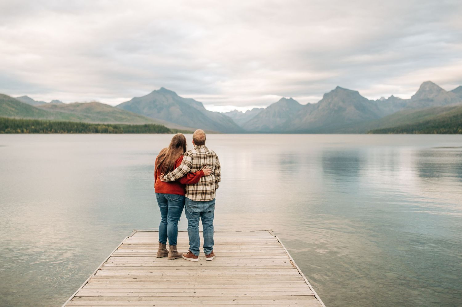 Two people stand together on a wooden dock overlooking a serene mountain lake with misty peaks in the distance.