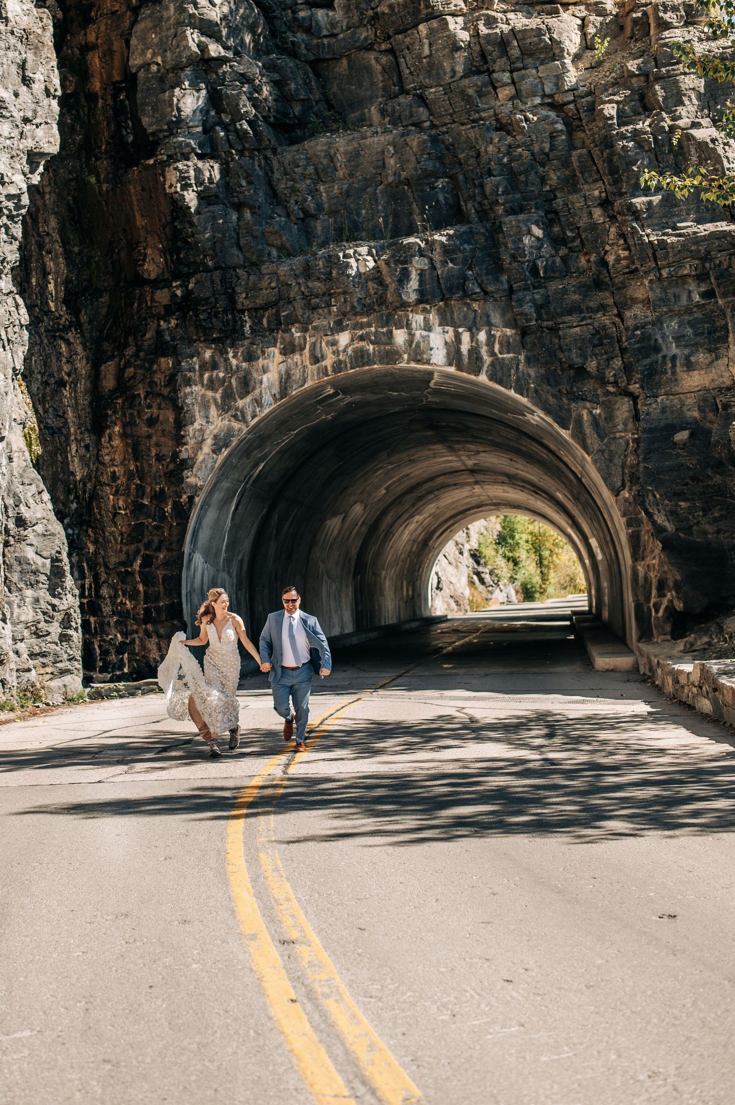 A stone tunnel carved through a mountain with sunlit road and two figures running through.