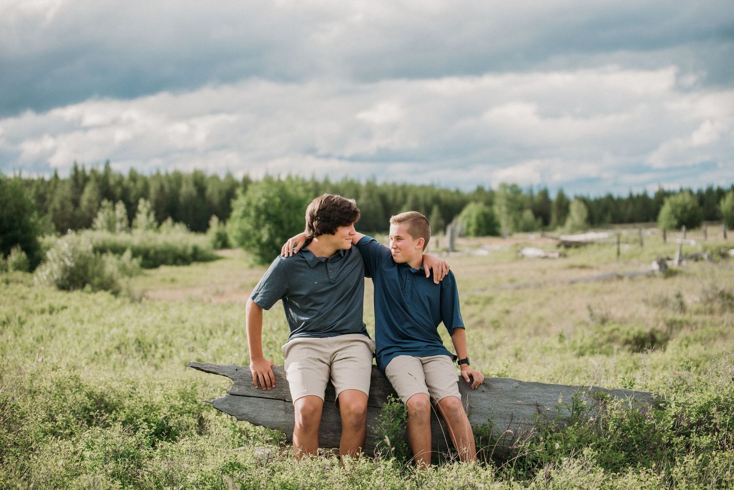 Two people sit close together on a fallen tree trunk in a scenic grassy meadow under cloudy skies.