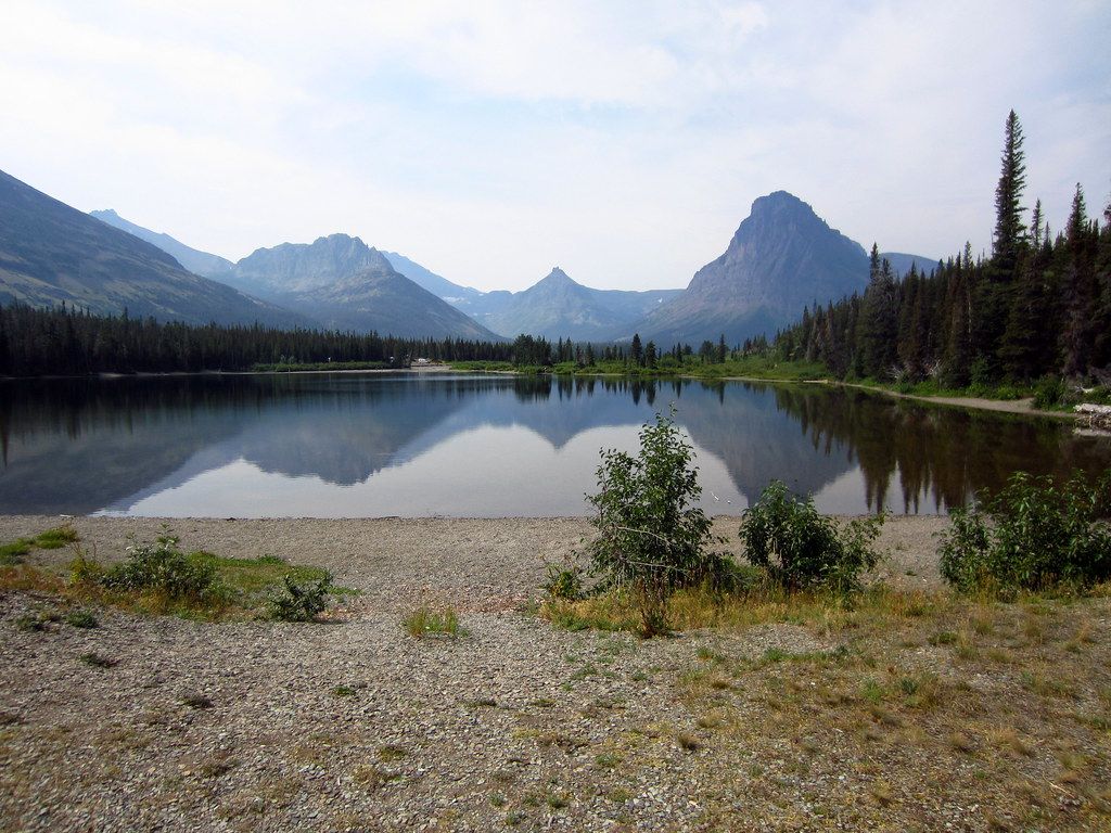 Tranquil mountain lake in Glacier National Park reflects jagged peaks and evergreen forest under clear blue sky.