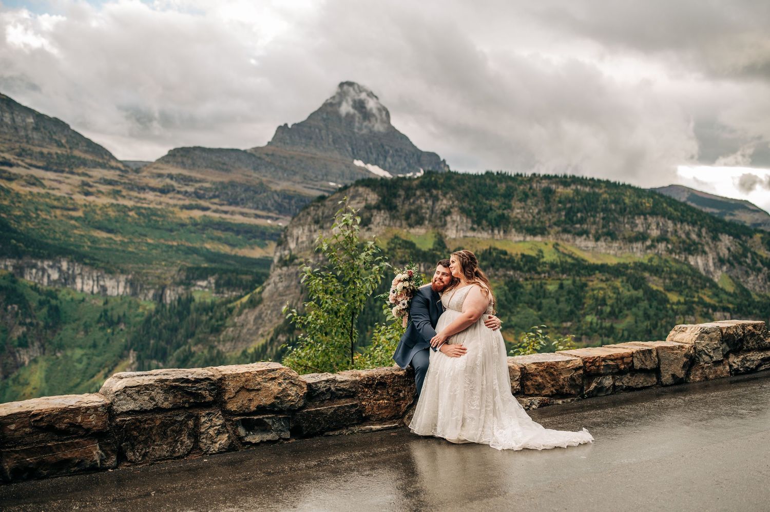 Two people share a romantic moment on Going-to-the-Sun Road in Glacier National Park with mountain peaks in the background.