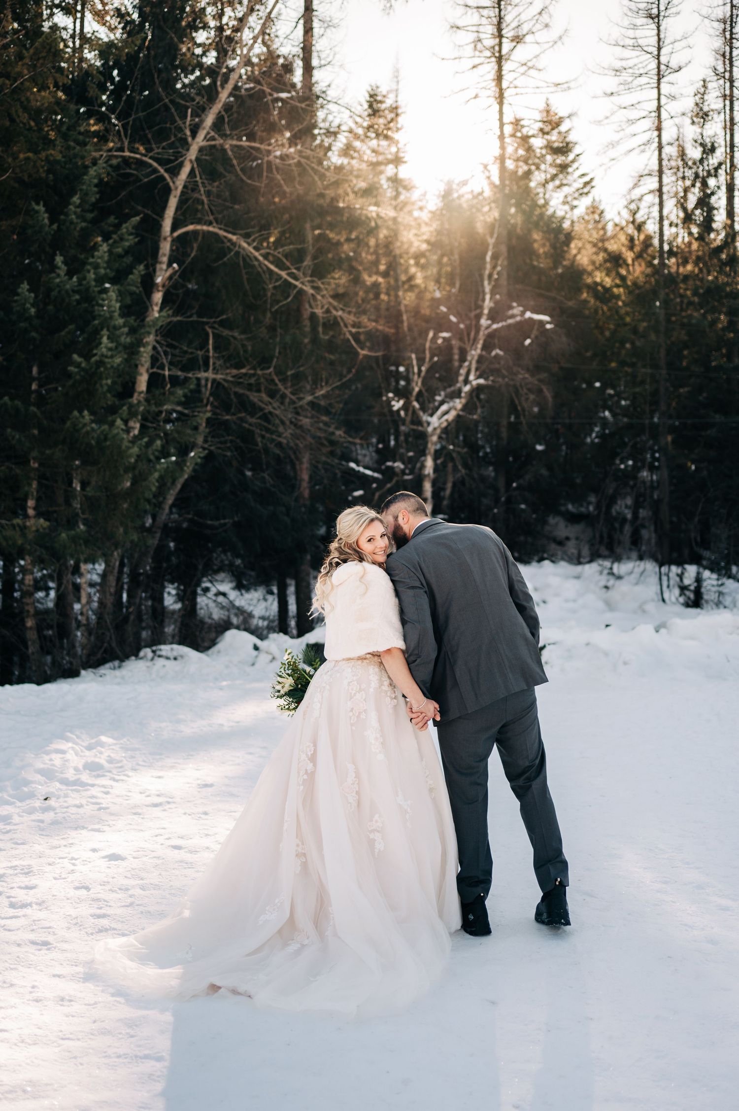Bride and groom holding hands in snowy winter forest at sunset, bride wearing white gown with fur wrap.
