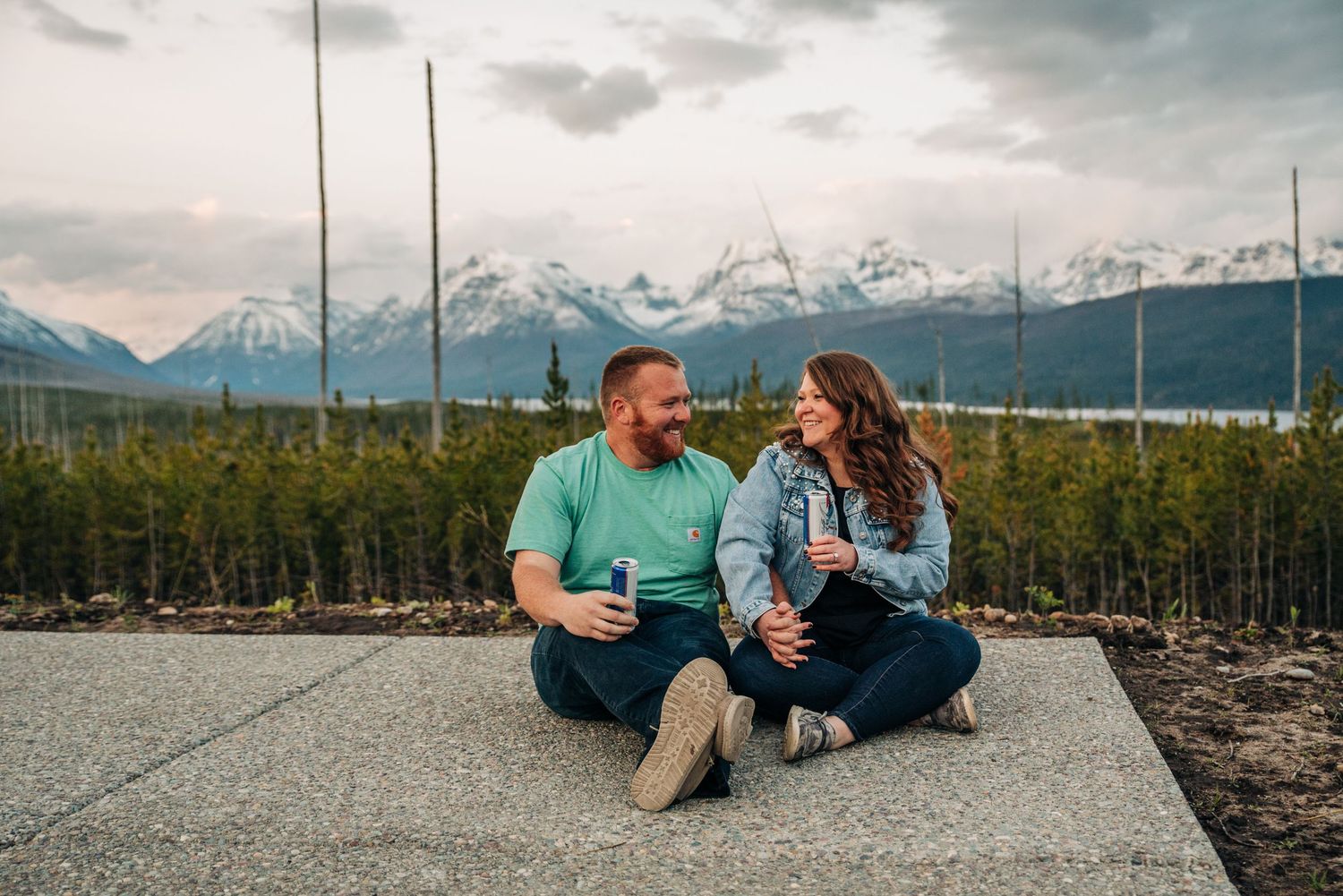 A couple sits together on a concrete slab with snow-capped mountains and evergreen trees in the background.