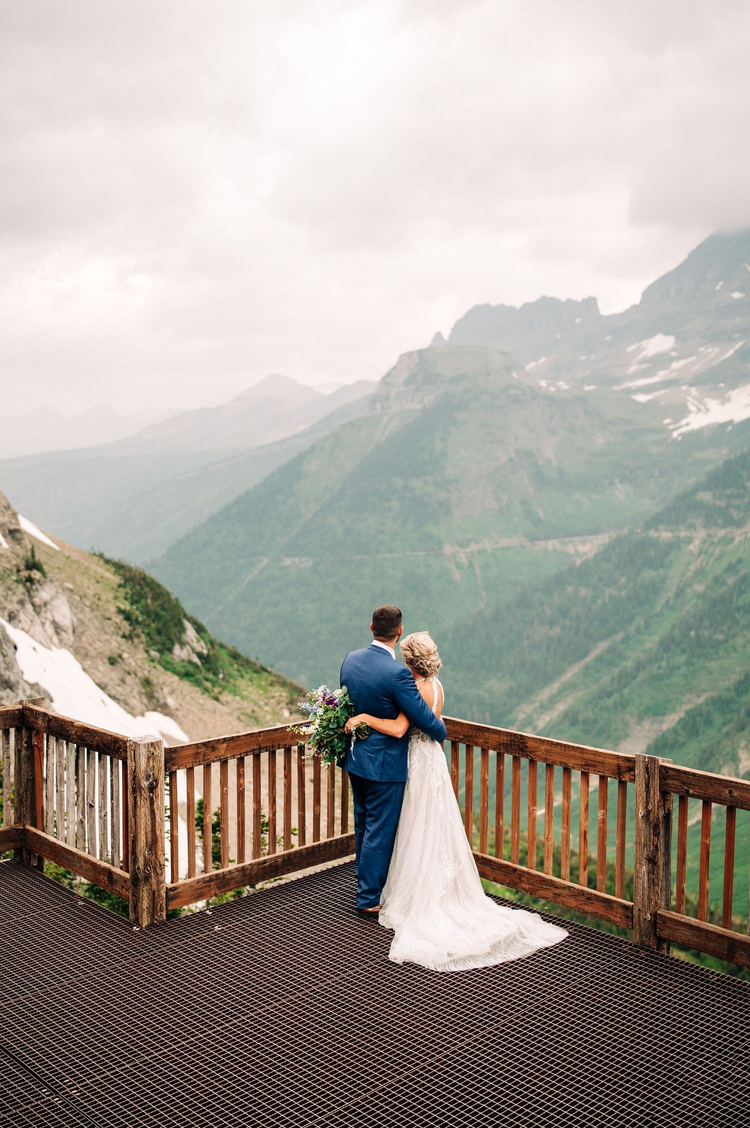 A couple embraces on a mountain observation deck overlooking snow-capped peaks in Glacier National Park.