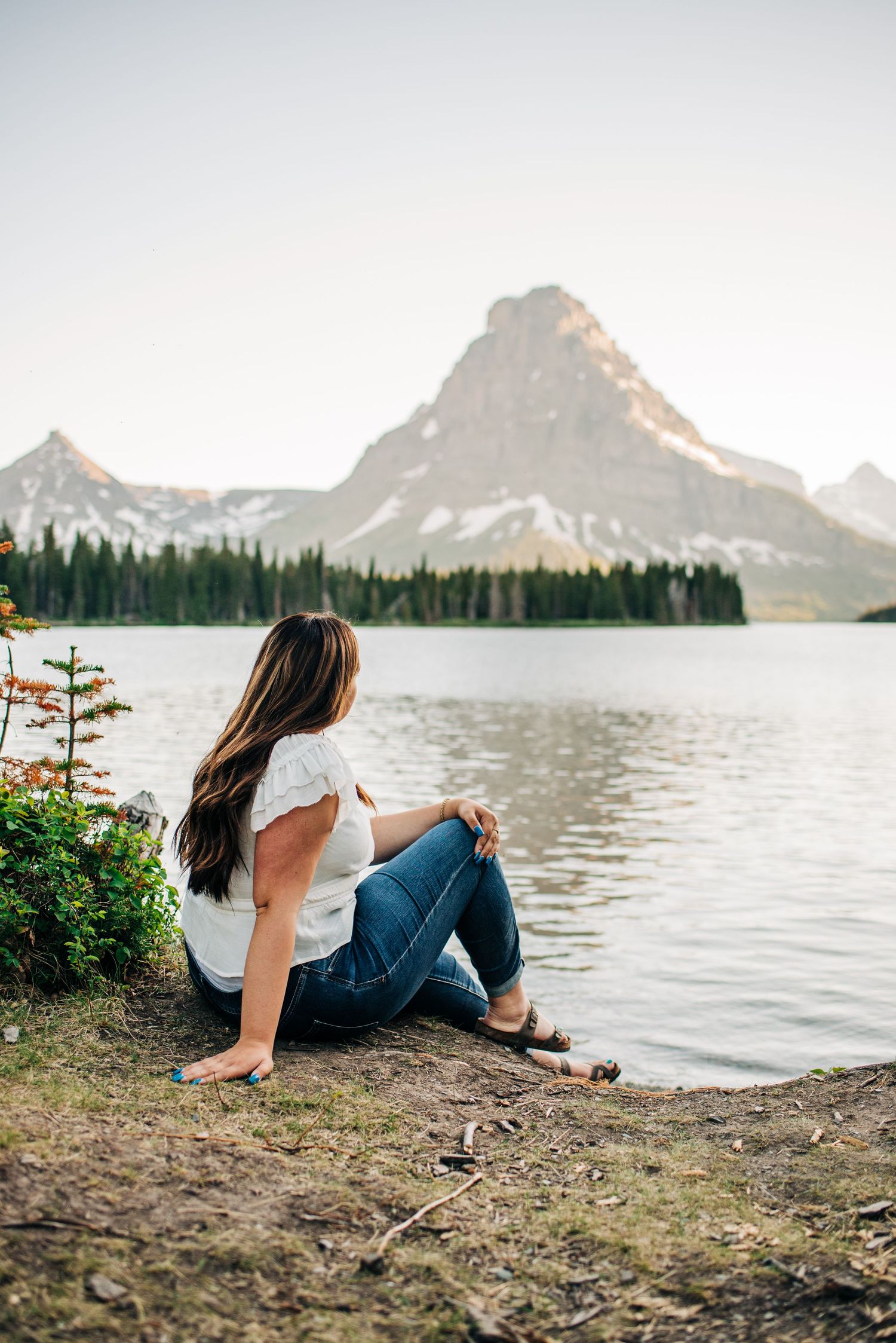 Person enjoying serene mountain lake view at sunset with evergreen trees and rugged peaks in background.