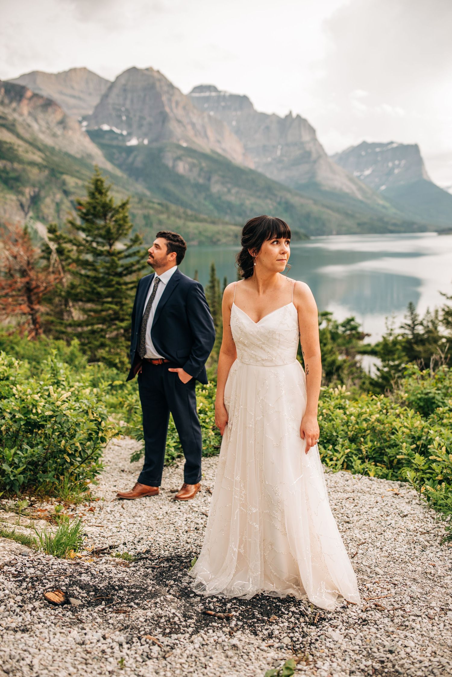 Wedding couple stands on rocky outlook with dramatic mountain lake and peaks creating stunning backdrop in nature.
